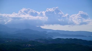 Duman yavaşça bacadan yükselerek yuvarlanan beyaz bulutlarla mavi gökyüzüne yükselir. Kıyı yeşil dağları sakin ve resimsel bir manzara yaratır. Jiufen, Tayvan.