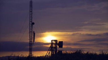 Radar istasyonu üzerinde parlak turuncu ve mor renkleri olan çarpıcı bir gün batımı. Siluetli iletişim kuleleri ve antenler renkli gökyüzüne karşı duruyor. Caoshan Radar İstasyonu, Tayvan.