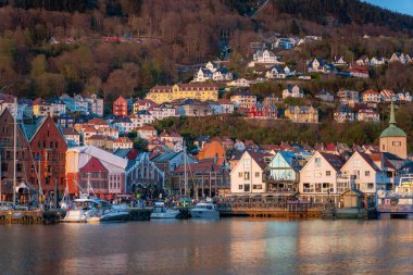 Bergen, Norveç - 30 Nisan 2024: Bergen, Norveç 'in renkli ahşap binalarıyla ikonik Bryggen rıhtımı insanlarla dolup taşıyor. Rıhtıma yanaşan tekneler büyüleyici manzarayı daha da güzelleştiriyor..