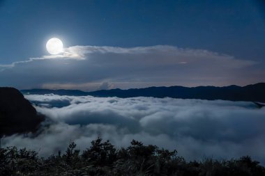 Dolunayın, okyanus dalgalarına benzeyen engin bir bulut denizini aydınlattığı sakin bir manzara. Gökyüzü yıldızlarla kaplı ve sakin bir ortam yaratıyor. Xindian Bölgesi, Tayvan.
