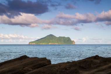 Kaplumbağa Adası 'nın denizdeki yemyeşil bitkilerle kaplı uzak manzarası. Beyaz bulutlar yavaşça yukarı doğru sürüklenerek adaya gölge düşürüyor. Şaşırtıcı doğal manzara, Yilan County, Tayvan.