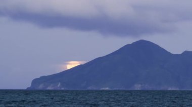 Dolunay yavaşça Guishan Adası 'nın arkasından yükseliyor. Parlak parlaklığı, geçen bulutlar tarafından kısmen gizleniyor. Sakin okyanus ruhani manzarayı yansıtır. Yilan County, Tayvan.