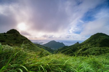 Uçsuz bucaksız mavi gökyüzünün altında yuvarlanan yeşil tepelerin ve uzak dağların panoramik görüntüsü. Okyanusa kısa bir bakış uzak mesafeden görülebilir. Jiufen, New Taipei Şehri, Tayvan.