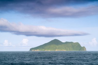 Ufukta Kaplumbağa Adası 'nın olduğu sakin bir kıyı manzarası. Ada engin bir mavi okyanus ve kabarık beyaz bulutlarla süslenmiş bir gökyüzü tarafından çerçevelenmiştir. Touchen, Yilan County, Tayvan.