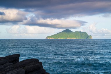 Ufukta Kaplumbağa Adası 'nın olduğu sakin bir kıyı manzarası. Ada engin bir mavi okyanus ve kabarık beyaz bulutlarla süslenmiş bir gökyüzü tarafından çerçevelenmiştir. Touchen, Yilan County, Tayvan.