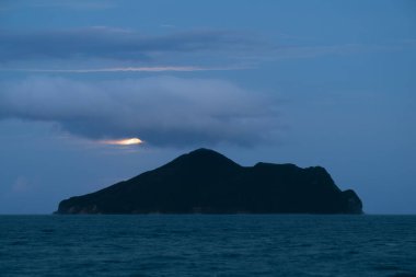 Dolunay yavaşça Guishan Adası 'nın arkasından yükseliyor. Parlak parlaklığı, geçen bulutlar tarafından kısmen gizleniyor. Sakin okyanus ruhani manzarayı yansıtır. Yilan County, Tayvan.