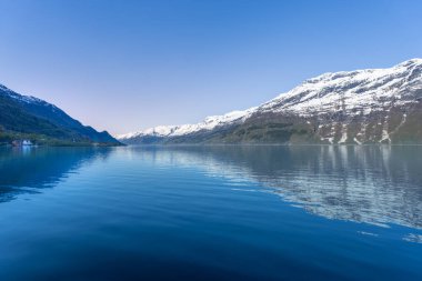 Hardangerfjord 'un sakin bir manzarası. Sakin turkuaz sular görkemli kar örtülü dağları ve berrak mavi gökyüzünü yansıtır. Huzurlu ve resimli bir sahne. Voss, Norveç.