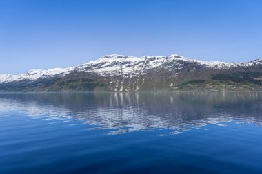 Hardangerfjord 'un sakin bir manzarası. Sakin turkuaz sular görkemli kar örtülü dağları ve berrak mavi gökyüzünü yansıtır. Huzurlu ve resimli bir sahne. Voss, Norveç.