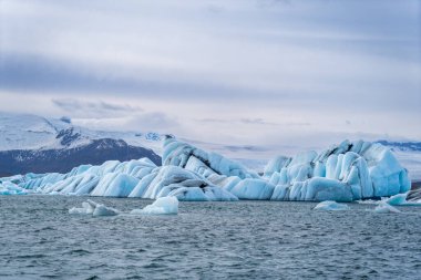 Blue and white icebergs float in a glacial lake against a backdrop of glaciers, creating a serene scene. Unspoiled Jokulsarlon, Iceland. clipart