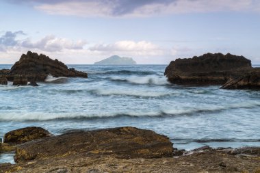 Gün batımında. Yumuşak dalgalar, sakin bir koyda yuvarlak kayalara çarpar. Gökyüzü durgun suyu yansıtan pembe ve mor yumuşak renklerle boyanmıştır. Toucheng, Yilan, Tayvan.
