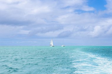Mauritius, Ile aux Cerfs 'in kristal berrak turkuaz sularında iki yelkenli süzülüyor. Nazik dalgalar teknelerin arkasında dalgalanma etkisi yaratıyor. Ile aux Cerfs, Mauritius, Hint Okyanusu.