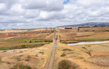 Scenic arched railway bridge crossing river in dry grassland with dramatic cloudy sky. Rural agricultural landscape with terraced fields and scattered trees, near Isalo National Park, Madagascar. clipart