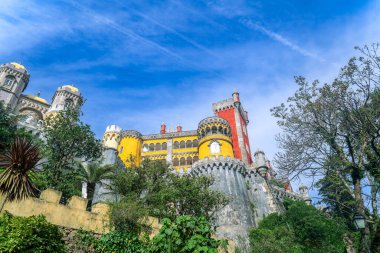 Pena Sarayı parlak sarı ve kırmızı kulelerini açık mavi gökyüzüne karşı sergiliyor. Yeşillik, tarihi yapıyı çevrelemektedir. Sintra, Portekiz.