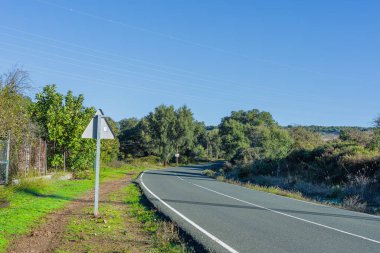 Yeşillik ve çeşitli bitki türleriyle çevrili dolambaçlı bir yol. Ek unsurlar arasında bir yol işareti ve elektrik hatları, Sierra de Aracena, İspanya.