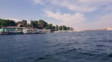 Nil Nehri kıyısı boyunca filikalar ve motorlu tekneler de dahil olmak üzere çeşitli tekneler demirlemiştir. Yeşil ağaçlar ve binalar kıyı boyunca uzanır, Aswan, Mısır 'da sakin bir manzara yaratır..