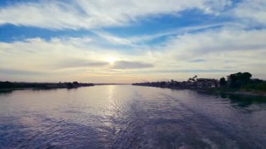 Mısır, Luxor yakınlarında mavi sularda parıldayan altın güneş ışığıyla nefes kesici bir Nil Nehri gün batımı. Bereketli yeşillik ve uzak palmiye ağaçları bir gemi gezisi sırasında çekilen sakin bir sahne oluşturur..