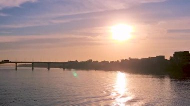 Mısır 'ın Luxor yakınlarındaki sakin mavi sulara yansıyan altın güneş ışığıyla huzurlu bir Nil Nehri gün batımı. Bir köprü ve uzak şehir manzarası. Alacakaranlıkta bir gemi gezisinde yakalanan cazibe..