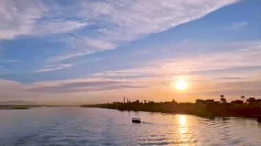 Mısır 'ın Luxor yakınlarındaki sakin mavi sulara yansıyan altın güneş ışığıyla çarpıcı bir Nil Nehri gün batımı. Palmiye ağaçları ve uzak bir şehir manzarasıyla çevrili küçük bir tekne huzur içinde süzülüyor..