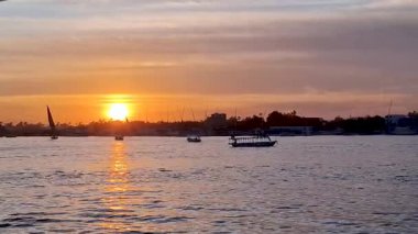 Güneş, sakin mavi Nil Nehri 'nin üzerinde batar ve suyun üzerine sıcak bir altın parıltı saçar. Teknelerin ve uzak binaların siluetleri canlı gökyüzüne karşı görünür. Luxor, Mısır.