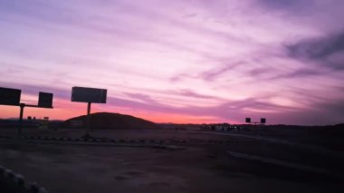 Alacakaranlıkta parlak pembe ve turuncu bir gökyüzü, karanlık dağ siluetleriyle, Luxor 'dan Hurghada, Mısır' a bir yol boyunca. Otoyoldaki araçlar sakin bir çöl manzarası yaratıyor..