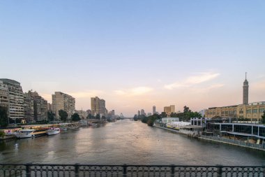 Kahire ufuk çizgisi alacakaranlıkta, Nil Nehri şehrin içinden akıyor. Nehir kıyısı boyunca yüksek binalar ve tekneler görülebilir. Gökyüzü mavi ve turuncu yumuşak renklerle boyanmıştır, Mısır.