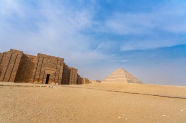 Açık mavi gökyüzünün altında kumlu bir çölde bej renkli bir Zoser Piramidi yükseliyor. Dikey desenli antik bir taş duvar ön planda duruyor, Saqqara, Mısır.