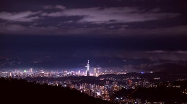 Bulutların, Taipei 'nin parıldayan silueti üzerinde büyüleyici bir dans edişini izleyin. Alçak bulutlar sola kayarken yüksek bulutlar sağa doğru süzülüyor, bahar gecesi gökyüzünün altında şiirsel bir tezat örüyor. Tayvan.