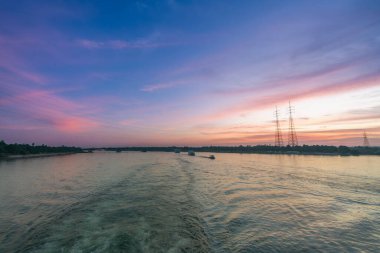 Pembe ve turuncu renkli canlı bir günbatımı Mısır 'ın Esna Kilidi yakınlarındaki Nil Nehri üzerinde gökyüzünü aydınlatıyor. Güç iletim kuleleri, bereketli palmiye ağaçları tarafından çerçevelenmiş nehir kıyısı boyunca dimdik dururlar..