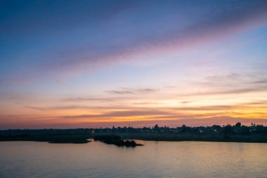 Esna Lock, Aswan, Mısır yakınlarındaki gün batımında Nil Nehri 'nin sakin geniş bir görüntüsü. Gökyüzü, uzak nehir kıyıları boyunca yumuşak pembe ve mor renkler, siluetli güç kuleleri ve palmiye ağaçları sergiliyor..
