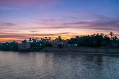 Mısır 'ın Aswan kentindeki Nil Nehri' nde büyüleyici bir alacakaranlık sahnesi, canlı pembe ve mor renkler gökyüzünü dolduruyor. Yerel evlerin ışıkları, palmiye ağaçlarının siluetleriyle nehir kıyısını aydınlatıyor..