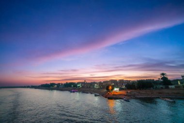 Mısır 'ın Aswan kentindeki Nil Nehri' nde büyüleyici bir alacakaranlık sahnesi, canlı pembe ve mor renkler gökyüzünü dolduruyor. Yerel evlerin ışıkları, palmiye ağaçlarının siluetleriyle nehir kıyısını aydınlatıyor..