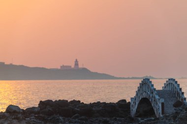 Tayvan 'ın New Taipei şehrindeki Shimen kıyı şeridi boyunca sakin bir günbatımı sahnesi, Fuguijiao Deniz feneri silueti yumuşak, sıcak gökyüzüne ve kıyı köprüsüne karşı..