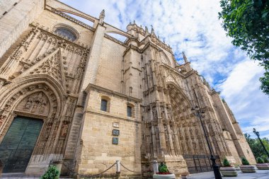 Bir UNESCO Dünya Mirası sahası olan Seville Katedrali 'nin karmaşık Gotik cephesinin düşük açılı görüntüsü, parçalı bulutlu bir gökyüzü altında devasa boyutlarını ve detaylı oymalarını sergiliyor, İspanya.