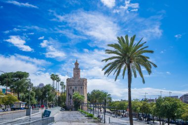 Sevilla, İspanya - 09 Mart 2025: Palmiye ağaçları ve mavi gökyüzü olan tarihi kule. İnsanlar yemyeşil asfalt yolda yürüyor. Flamenko mirası ve kültür alanlarıyla tanınan.