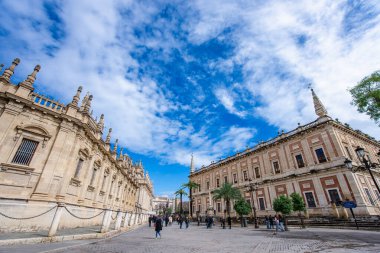 Sevilla, İspanya - 10 Mart 2025: Süslü cepheli ve palmiye ağaçlarıyla dolu tarihi binalar, dağınık bulutlu canlı mavi bir gökyüzünün altında hareketli bir cadde. İnsanlar bölgeyi gezip keşfediyorlar..