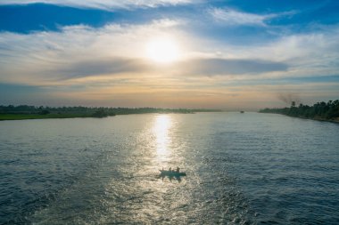 Güneş Nil Nehri 'nin üzerinde batar ve suyun yüzeyinde altın bir yansıma yaratır. Nehrin ortasında içinde insanlar olan küçük bir balıkçı teknesi görülüyor ve her iki yakasında yemyeşil bankalar var..