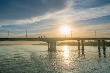 Güneş, Luxor yakınlarındaki Nil Nehri üzerindeki bir köprünün üzerinde batıyor ve ışığı suya yansıyor. Şehir manzarası uzaktan görülebiliyor, güzel manzarayı tamamlıyor..