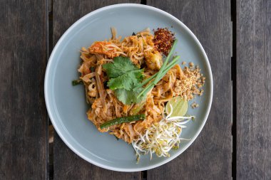 Top down view of a delicious pad thai dish (Thai stir fried noodles) on a blue plate with chilli flakes, peanuts, lime and beansprouts