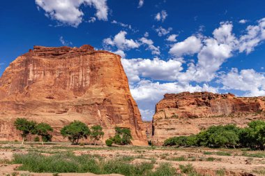 Arizona 'daki Chelly Ulusal Anıtı Kanyonu' ndaki bulutlu mavi gökyüzü altındaki kırmızı kaya kayalıklarının ve yeşil ağaçların çarpıcı manzarası..