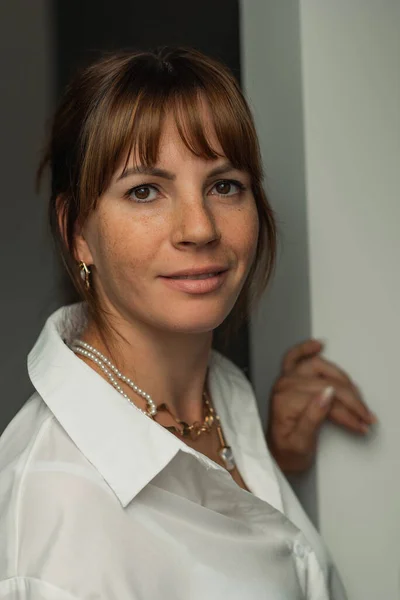 stock image A white woman with freckles in a white shirt. Aesthetic photo, calm emotions, standing against the wall