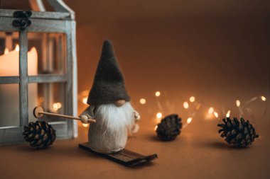 Güzel bir cüce, uzun şapkalı, kayak takımında ve kahverengi arka planda, mum, koni ve çelenkle süslenmiş. Kartpostallara ve Noel kutlamalarına uygun.