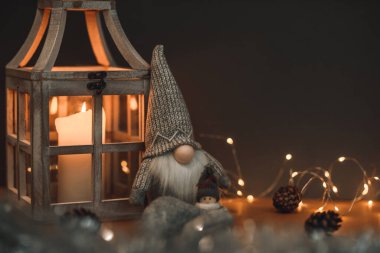 Uzun şapkalı sevimli bir cüce ve kahverengi arka planda bir kardan adam, mum, koni ve çelenkle süslenmiş. Kartpostallara ve Noel kutlamalarına uygun.