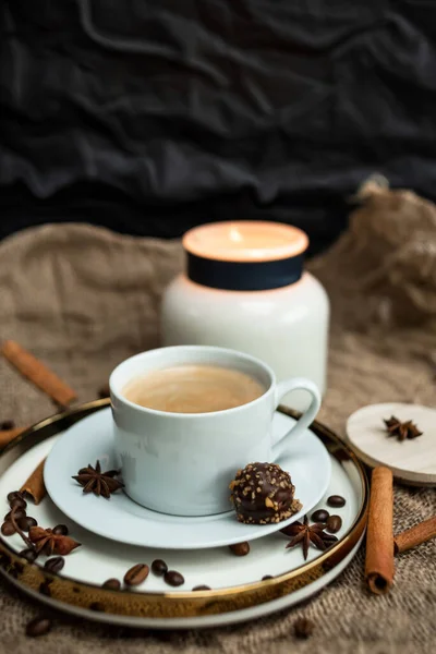 An aesthetic composition of a cup of black coffee with foam and a burning candle, decorated with chocolates, anise stars, coffee beans and cinnamon sticks
