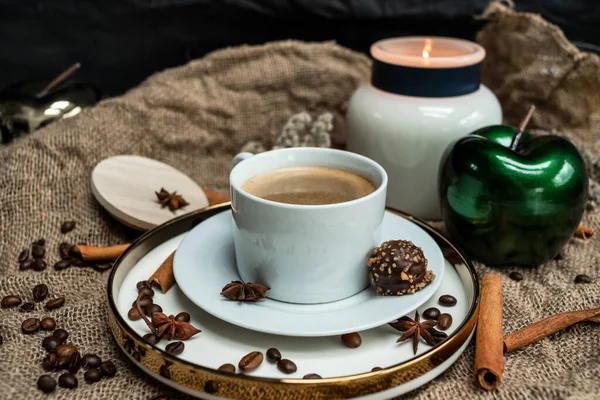 An aesthetic composition of a cup of black coffee with foam and a burning candle, decorated with chocolates, anise stars, coffee beans and cinnamon sticks