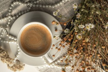 A white cup of coffee on a saucer, with a string of pearls, dried flowers, a magazine and a newspaper. Aesthetic picture, elegant background photo.