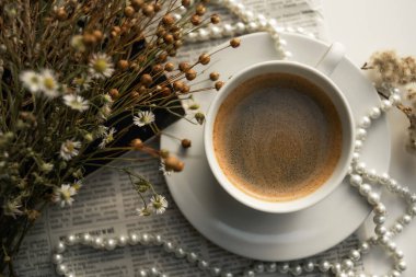 A white cup of coffee on a saucer, with a string of pearls, dried flowers, a magazine and a newspaper. Aesthetic picture, elegant background photo.