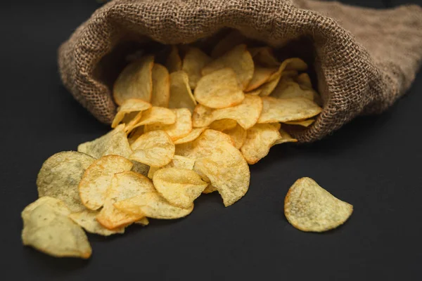 A canvas bag with potato chips scattered across a black table. Potato chips spilled out of bag