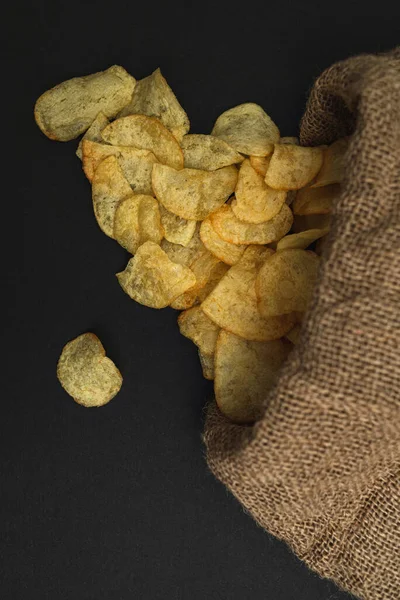 A canvas bag with potato chips scattered across a black table. Potato chips spilled out of bag