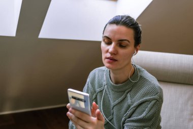 Kulaklık takan, orta yaşlı bir kadın sandalyede oturuyor ve akıllı telefondan video görüşmesi yapıyor.