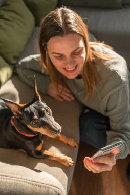 Kanepede bir köpek kameraya bakıyor, bir kadın telefona bakıyor, kanepenin ve köpeğin yanında oturuyor.